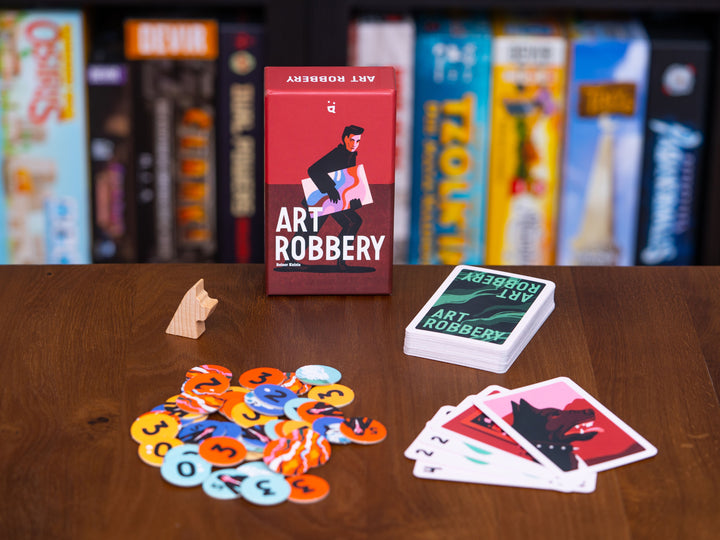 A photo of the box, cards, cardboard tokens, and single wooden token included in the board game Art Robbery, against a backdrop of other board games on shelves. The cover displays the name of the game in white, against a red background, and a figure in black holding a piece of art with swirly colors. Some cards are red, while the majority are stacked in a deck with a swirly green background. The cardboard tokens are a variety of colors with a single number printed in the center.