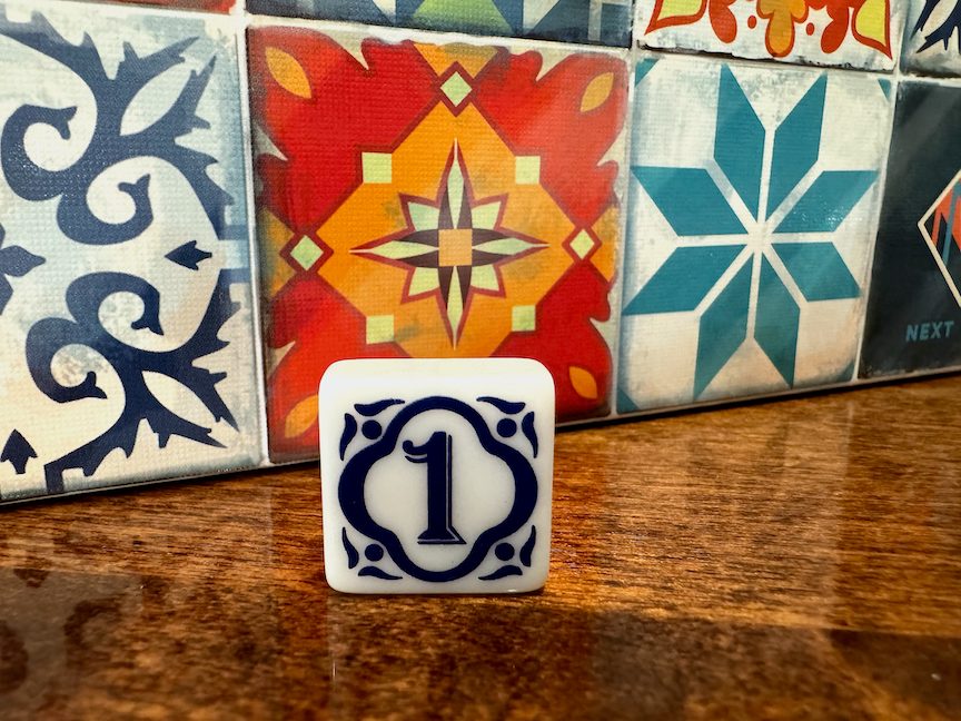 A close up photo of the 1st Player tile for use with the board game Azul. The tile is sitting on a polished wooden surface and the game box is partially visible behind it. The tile is white with a blue engraved pattern with the number 1 in the center.