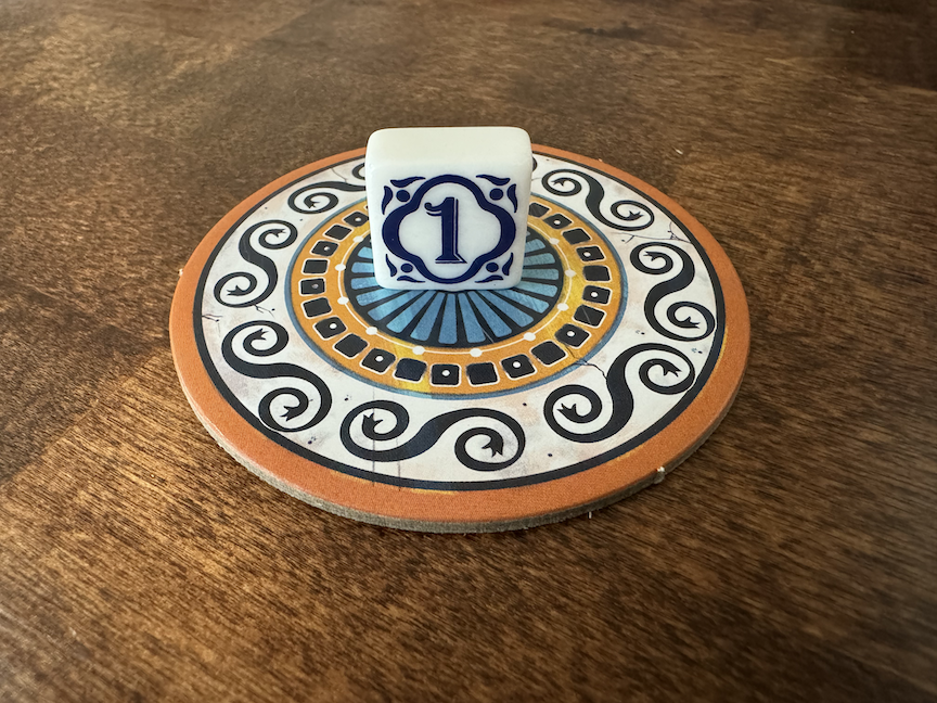A close up photo of the 1st Player tile for use with the board game Azul. The tile is sitting on a circular cardboard piece from the board game on a polished wooden surface. The tile is white with a blue engraved pattern with the number 1 in the center.