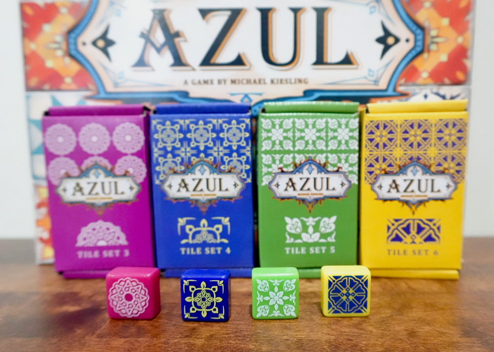 A photo of the 3rd, 4th, 5th, and 6th tile sets for use with the board game Azul. The four boxes are sitting in front of the game box and have one example tile sitting in front of the tile set boxes.