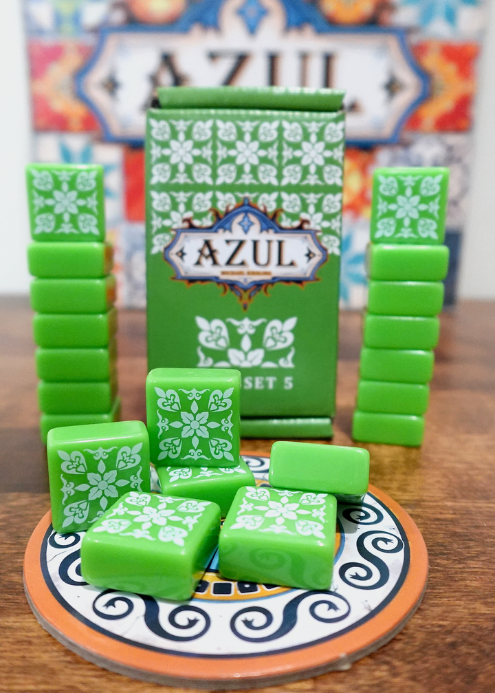 A photo of the fifth tile set for use with the board game Azul. These square tiles are green with a white pattern printed on one side. A pile is on top of one of the cardboard pieces from the game, with the box and remaining tiles stacked in the back.