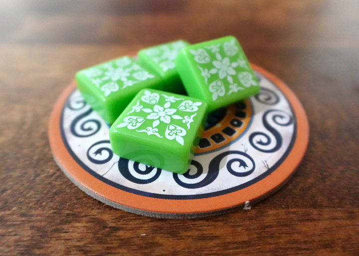 A photo of the fifth tile set for use with the board game Azul. These square tiles are green with a white pattern printed on one side. A small pile of tiles is sitting on a circular cardboard piece from the game, which in turn is on a polished wooden surface