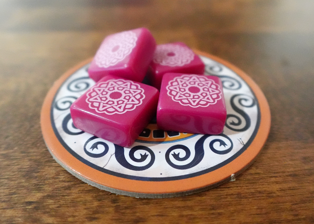 A photo of the third tile set for use with the board game Azul. These square tiles are magenta with a white pattern printed on one side. A small pile of tiles is sitting on a circular cardboard piece from the game, which in turn is on a polished wooden surface.