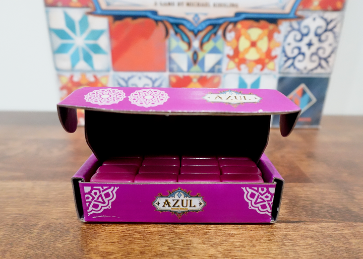 A photo of the third tile set for use with the board game Azul. These magenta tiles are sitting in a matching magenta box, which in turn is sitting in front of the game box of Azul.