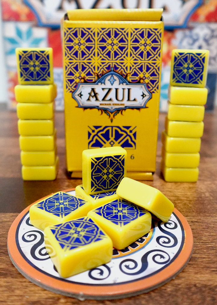 A photo of the sixth tile set for use with the board game Azul. These square tiles are yellow with a dark blue pattern printed on one side. A pile is on top of one of the cardboard pieces from the game, with the box and remaining tiles stacked in the back.