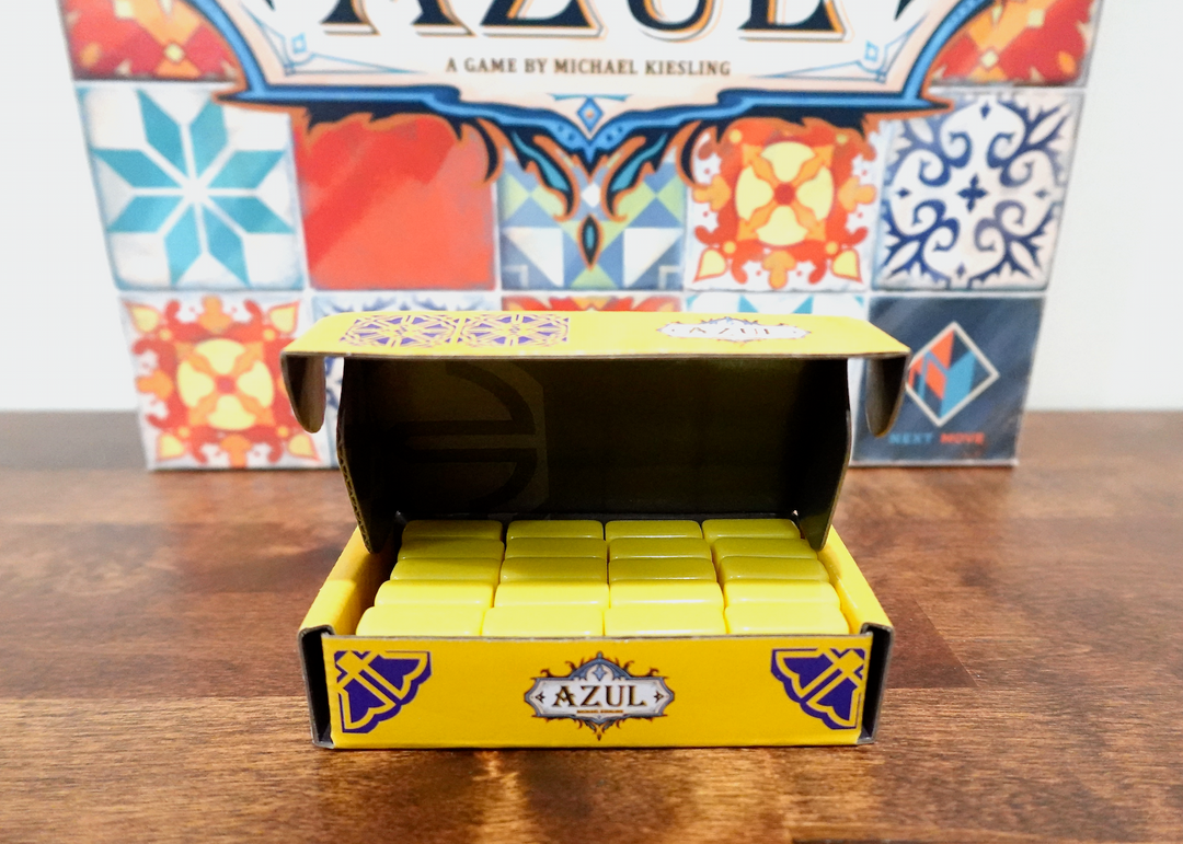 A photo of the sixth tile set for use with the board game Azul. These yellow tiles are sitting in a matching yellow box, which in turn is sitting in front of the game box of Azul.
