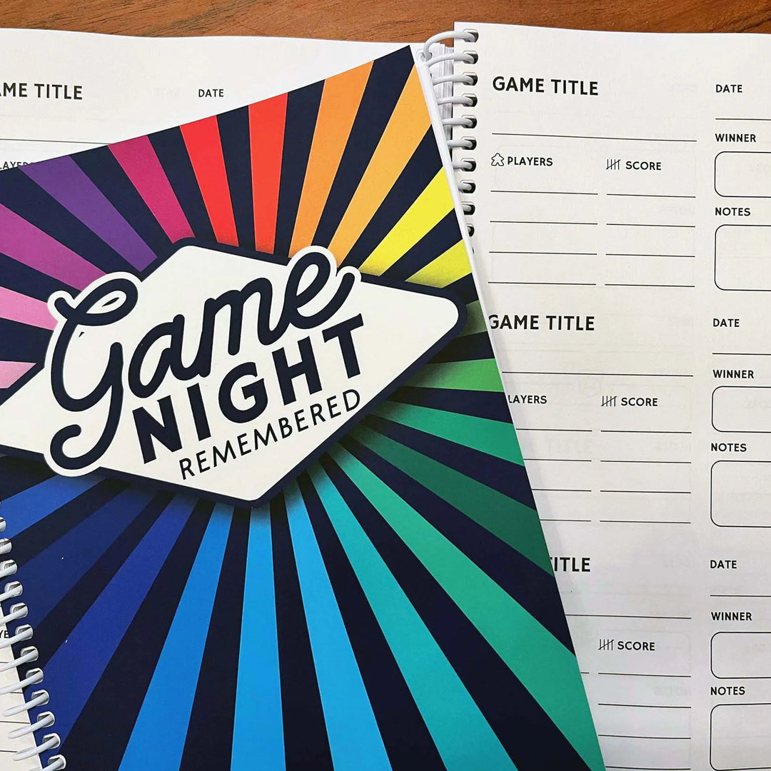 A spiral bound notebook sitting on top of open pages. The cover says "Game Night Remembered" on a rainbow starburst background. The open pages in the background have sections to record aspects of playing board games.