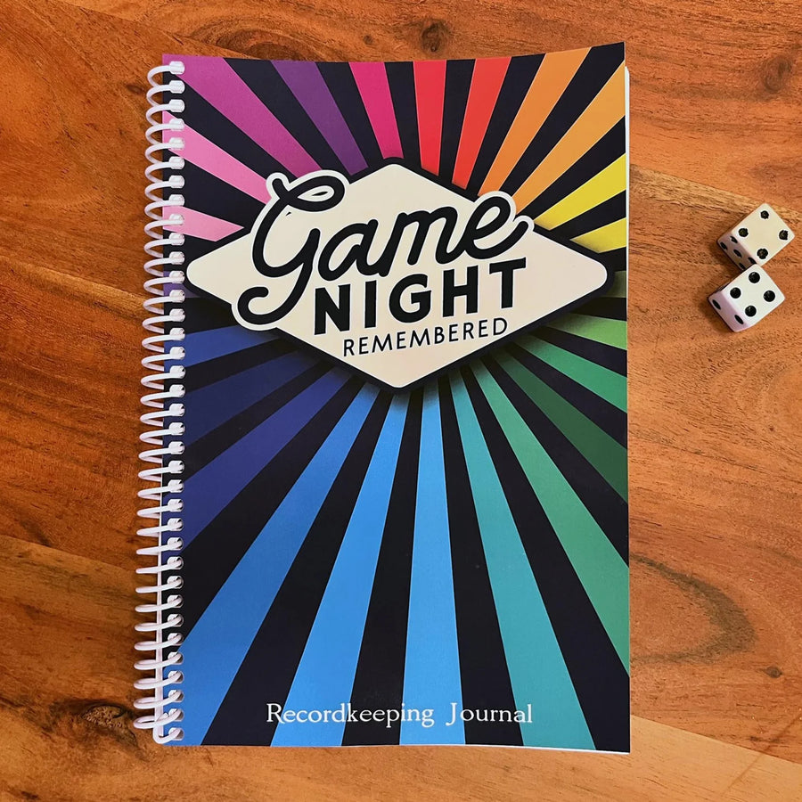 A spiral bound notebook sitting next to a pair of dice on a wooden tabletop. The cover says "Game Night Remembered" on a rainbow starburst background.