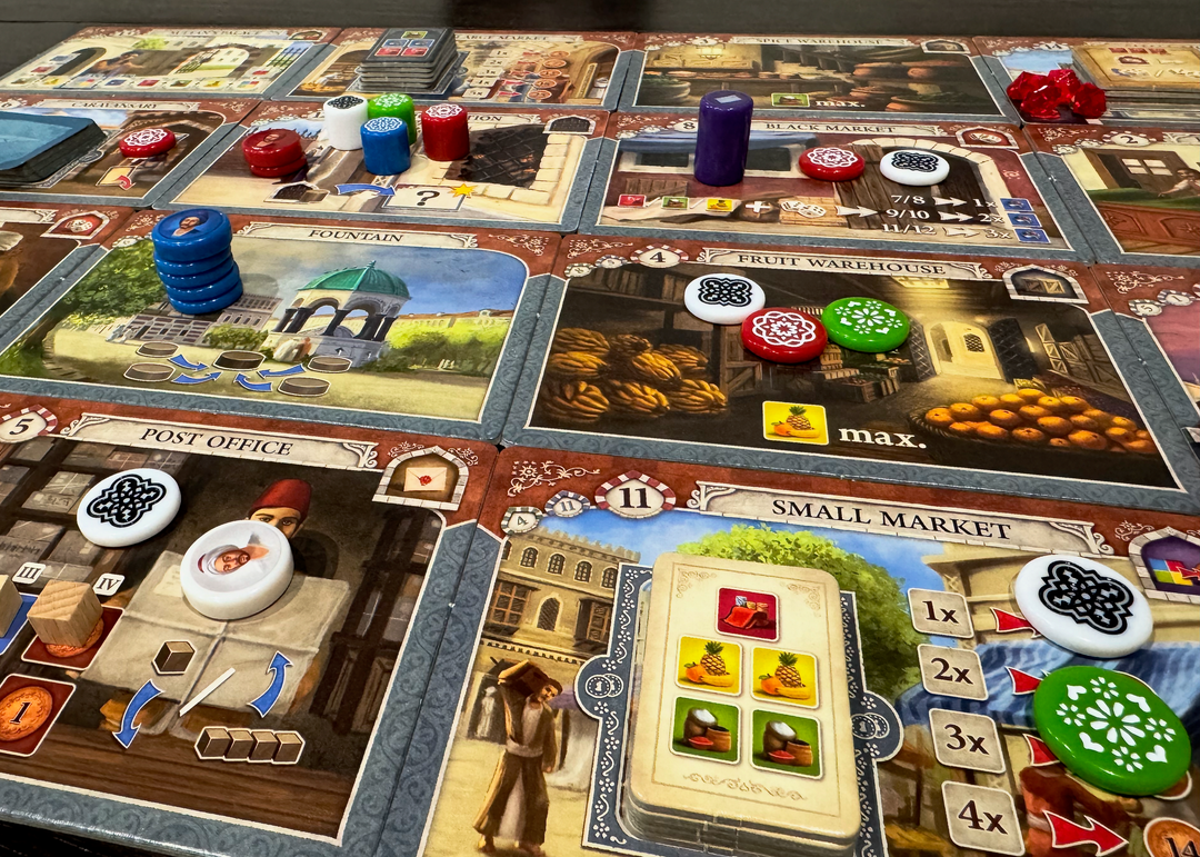A photo of the game board from the board game Istanbul, featuring cardboard rectangles laid out in a grid, each with a different illustration and symbols relavant to gameplay. On top of some of these rectangles are engraved plastic pieces in a variety of colors, heights, and diameters.