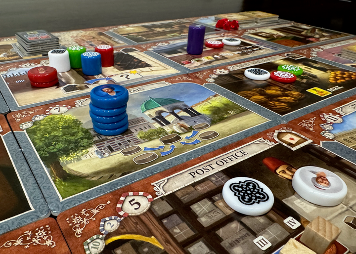 A photo of the game board from the board game Istanbul, featuring cardboard rectangles laid out in a grid, each with a different illustration and symbols relavant to gameplay. On top of some of these rectangles are engraved plastic pieces in a variety of colors, heights, and diameters.