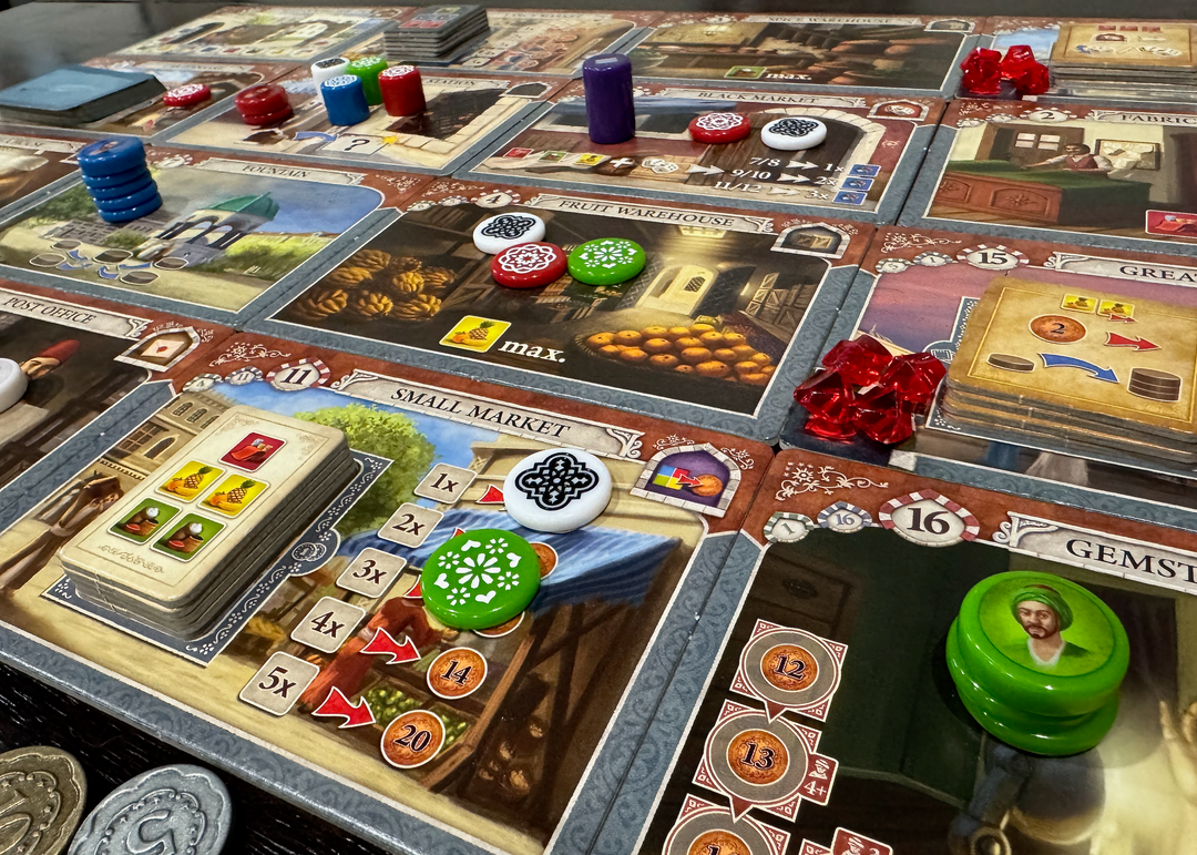 A photo of the game board from the board game Istanbul, featuring cardboard rectangles laid out in a grid, each with a different illustration and symbols relavant to gameplay. On top of some of these rectangles are engraved plastic pieces in a variety of colors, heights, and diameters.
