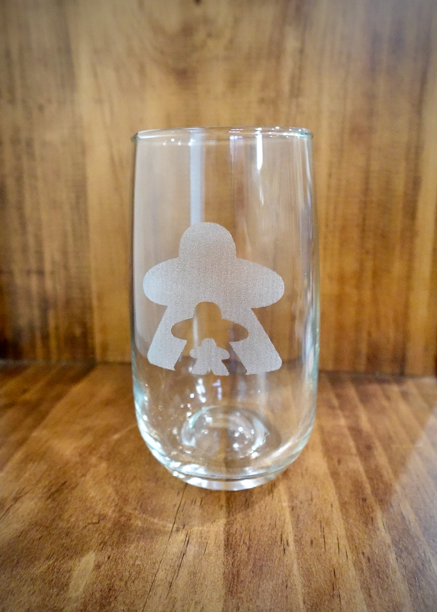 A photo of a drinking glass against a wooden background. The drinking glass features a frosted design of three person-shapes of different sizes.