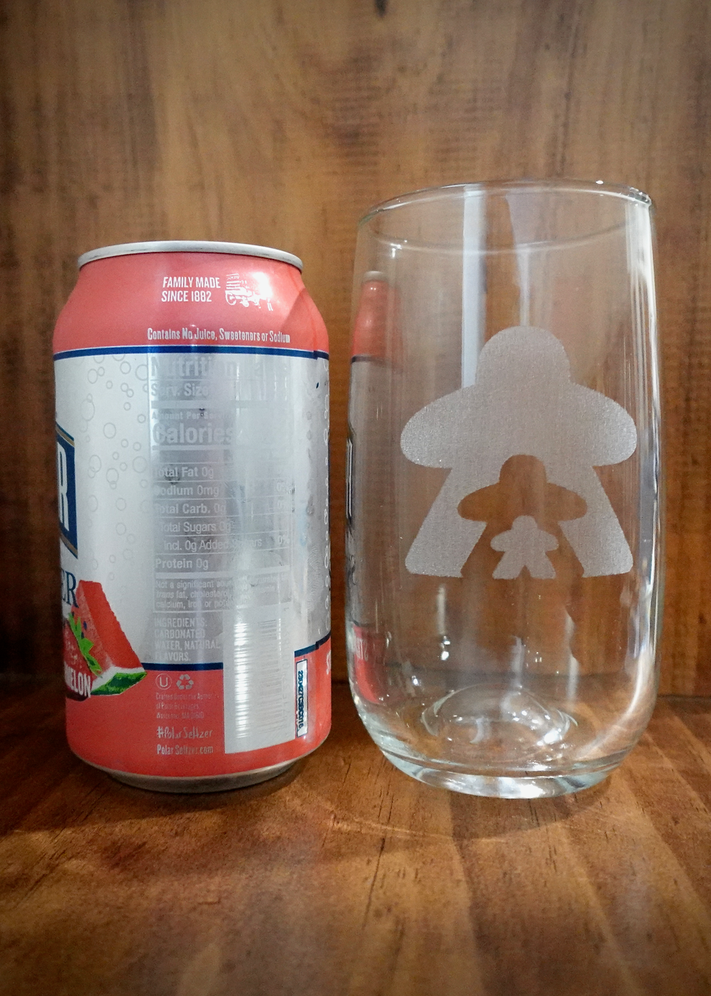 A photo of a drinking glass next to a soda can against a wooden background. The drinking glass features a frosted design of three person-shapes of different sizes.