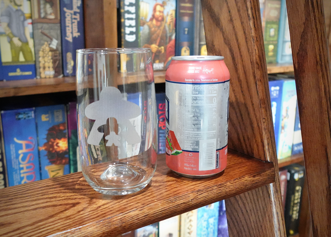 A photo of a drinking glass next to a soda can, sitting on the step of a ladder in front of a wall of board game boxes. The drinking glass features a frosted design of three person-shapes of different sizes.
