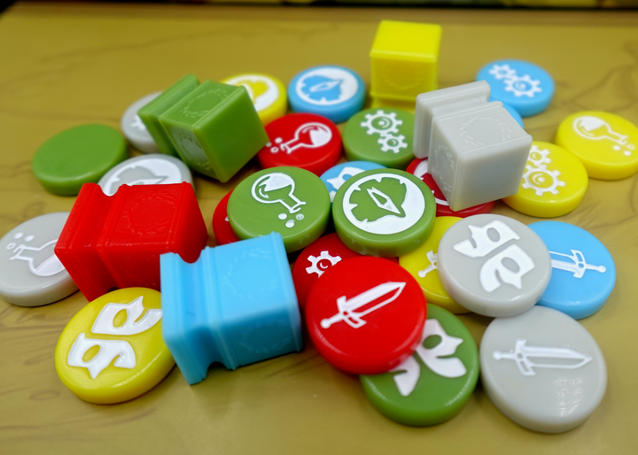 A side-view photo of a jumble of GeekUp bits for use with the board game Tapestry. Most bits are circular disks in a variety of colors, each with an engraved symbol in the center. The rest of the tokens are square, plastic columns with a laurel wreath printed on each side.