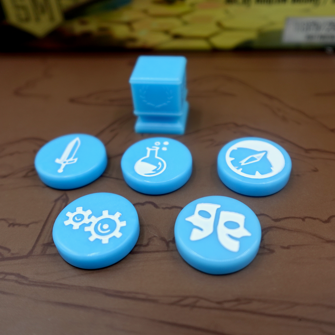 A close up photo of the blue GeekUp tokens for use with the board game Tapestry.  Five of the bits are circular disks, each with an engraved symbol in the center. The remaining token is a square, plastic column with a laurel wreath printed on each side.