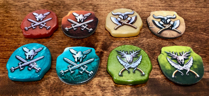 A photo of plastic upgraded tokens for use with the board game Tash-Kalar, sitting next to a matching cardboard tokens of the same shape and color. Each of the tokens, both plastic and cardboard, has a different type of weapon printed on the top.
