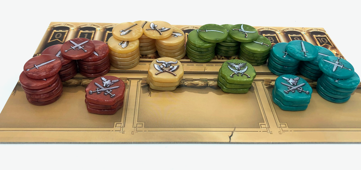 A photo of plastic upgraded tokens for use with the board game Tash-Kalar, sitting in piles by color: red, yellow, green, and blue, on one of the boards from the game. Each of the plastic tokens has a different type of weapon printed on the top.