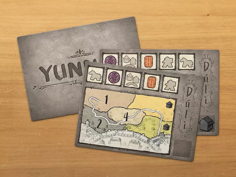 A photo of some components from the board game Yunnan. Pictured are three cards on a wooden background: one with the game's titles, and two with symbols and a map.