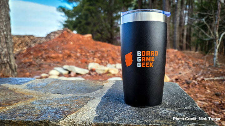 A black and silver travel mug, with a clear, push top above the silver rim, sitting outside on a stone wall with a forest in the background. The mug is decorated with an orange logo and the words "BoardGameGeek".