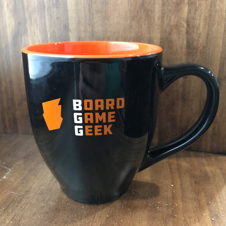 A bistro-shaped, glossy black coffee mug, with orange on the inside, labeled with "BoardGameGeek" on the outside, against a polished wooden background.