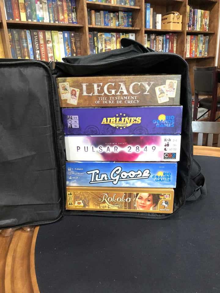 A black carry bag and backpack, sitting on a padded tabletop, with five board games stacked vertically inside, and multiple bookcases of board games in the background.
