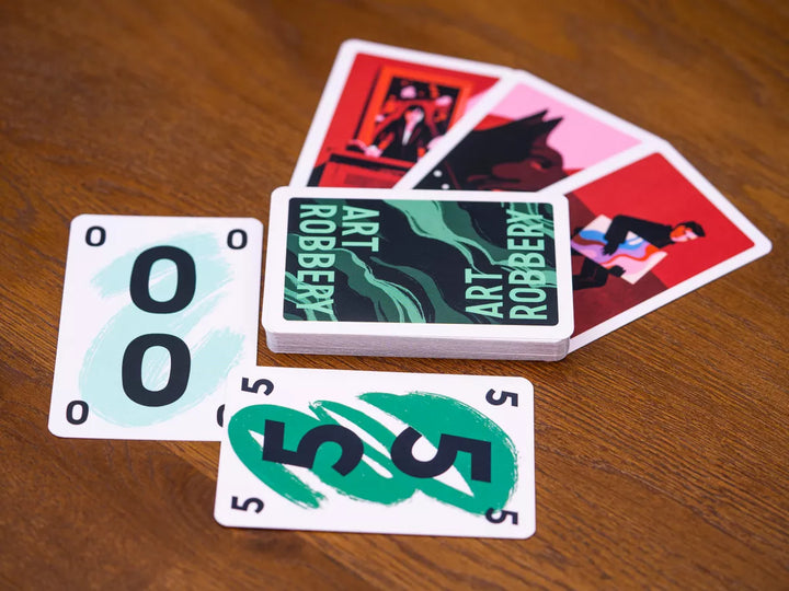 A photo of the cards from the board game Art Robbery, pictured on a wooden tabletop. Three of the cards in the background feature red arwork. The rest of the cards display a swirly green background on the backs, and numbers in both the center and corners on the front.