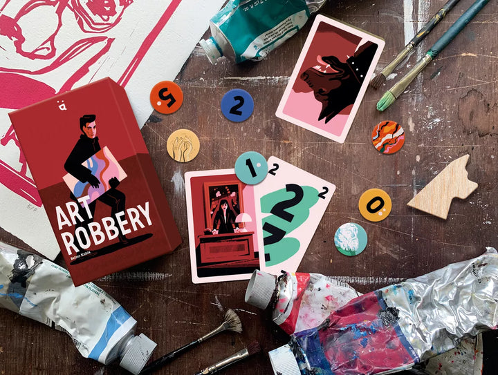 A photo of the box and components from the board game Art Robbery, on a battered wooden tabletop, surrounded by used paint tubes, canvas, and paint brushes,