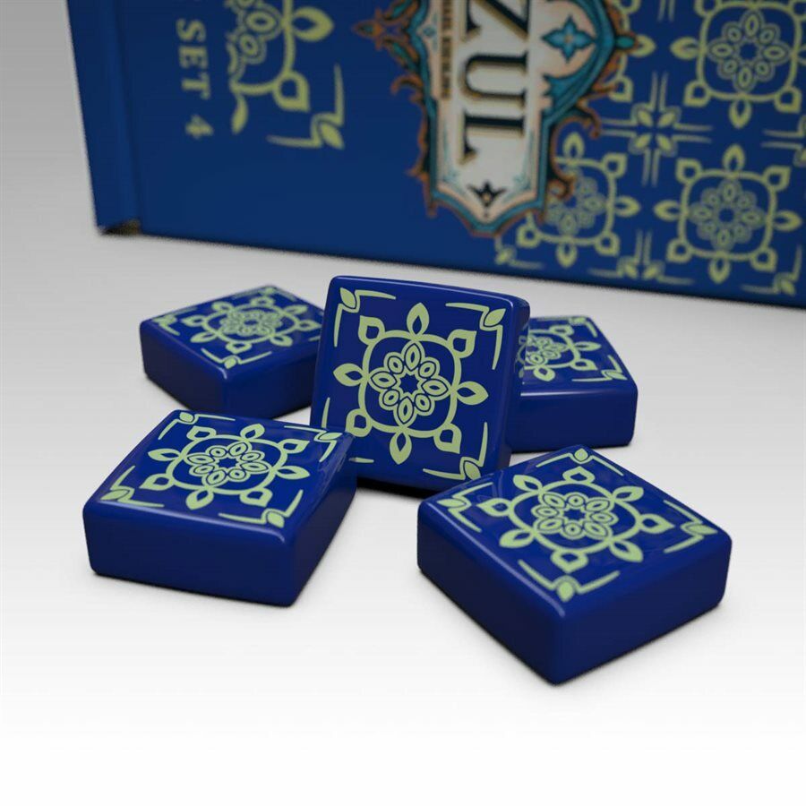 A photo of the third tile set for use with the board game Azul. These square tiles are dark blue with a yellow pattern printed on one side. A pile of tiles is sitting on a white background with the box partially visible in the back.