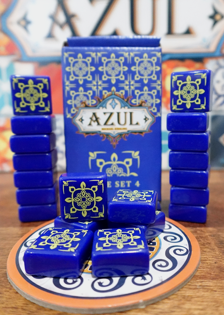 A photo of the fourth tile set for use with the board game Azul. These square tiles are dark blue with a yellow pattern printed on one side. A pile of tiles is on top of one of the cardboard pieces from the game, with the box and remaining tiles stacked in the back.