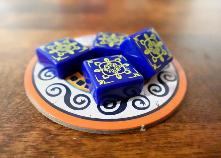 A photo of the fourth tile set for use with the board game Azul. These square tiles are dark blue with a yellow pattern printed on one side. A small pile of tiles is sitting on a circular cardboard piece from the game, which in turn is on a polished wooden surface