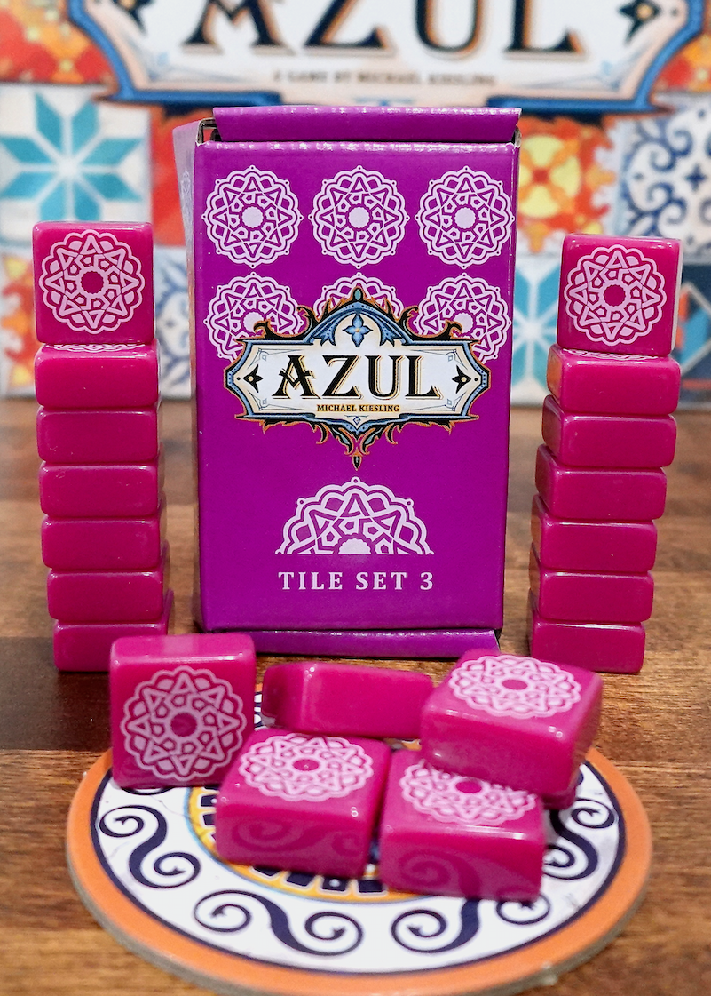 A photo of the third tile set for use with the board game Azul. These square tiles are magenta with a white pattern printed on one side. A pile is on top of one of the cardboard pieces from the game, with the box and remaining tiles stacked in the back.