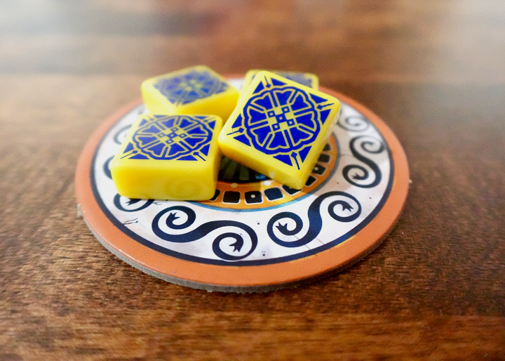 A photo of the sixth tile set for use with the board game Azul. These square tiles are yellow with a dark blue pattern printed on one side. A small pile of tiles is sitting on a circular cardboard piece from the game, which in turn is on a polished wooden surface.
