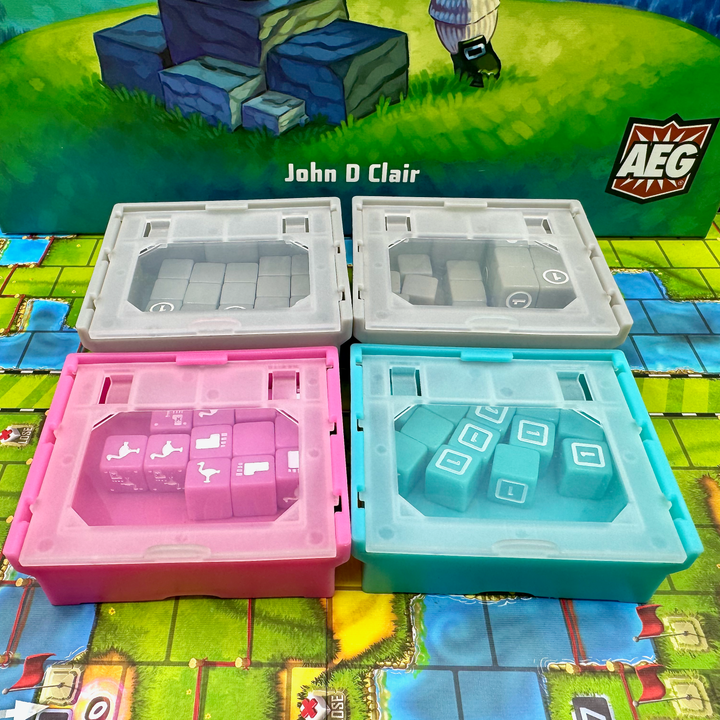 A photo of four, plastic storage boxes for use with the board game Cubitos. Each box has a clear top with dice of a matching color inside. One box is pink, one teal, and two gray. The boxes are sitting on the game board from Cubitos and part of the image of the game box cover - a foot and cube-shaped blocks in a grassy field - is visible in the background.