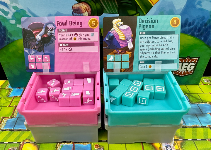 A photo of four, plastic storage boxes for use with the board game Cubitos. Each box has a clear top with dice of a matching color inside. One box is pink, one teal, and two gray. The pink and teal boxes are sitting on top of the gray boxes and have their lids flipped open. A card from the game is resting on each lid. The boxes are sitting on the game board from Cubitos and part of the image of the game box cover is visible in the background.