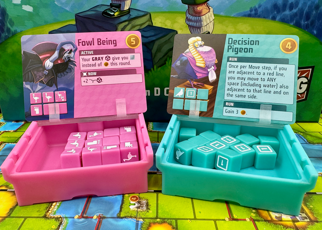 A photo of two, plastic storage boxes for use with the board game Cubitos. One box is pink and the other one teal. Both boxes have their lids flipped open. A card from the game is resting on each lid. The boxes are sitting on the game board from Cubitos and part of the image of the game box cover is visible in the background.