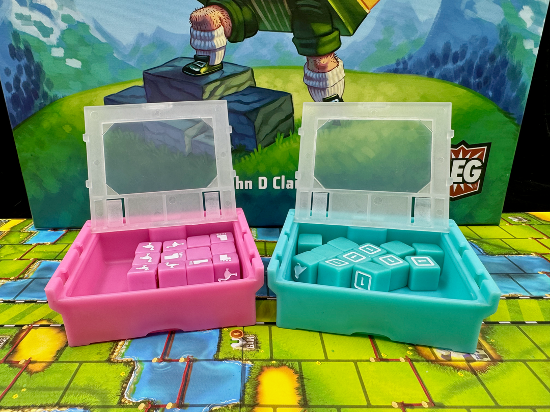 A photo of two, plastic storage boxes for use with the board game Cubitos. One box is pink and the other one teal. Both boxes have their lids flipped open. A card from the game is resting on each lid. The boxes are sitting on the game board from Cubitos and part of the image of the game box cover is visible in the background.