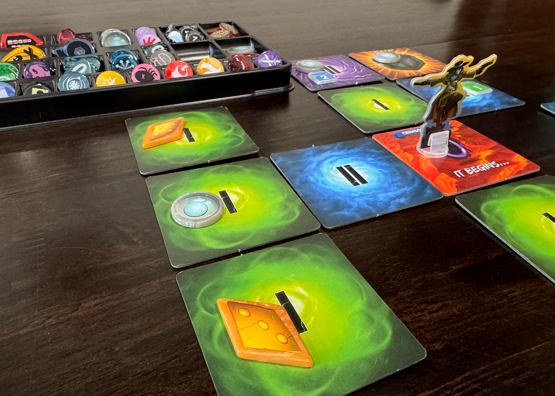 A photo of some of the original and upgraded pieces from the board game Dice Throne Adventures, pictured on a dark, wooden tabletop. Visible in the foreground are a grid of square, cardboard tiles each color coded green, blue, or purple and with a Roman numeral. On top of some of those tiles are plastic, GeekUp tokens , with artwork from the game printed on the top. In the background, is a black, plastic tray with compartments that hold all of the remaining GeekUp bits in this set.