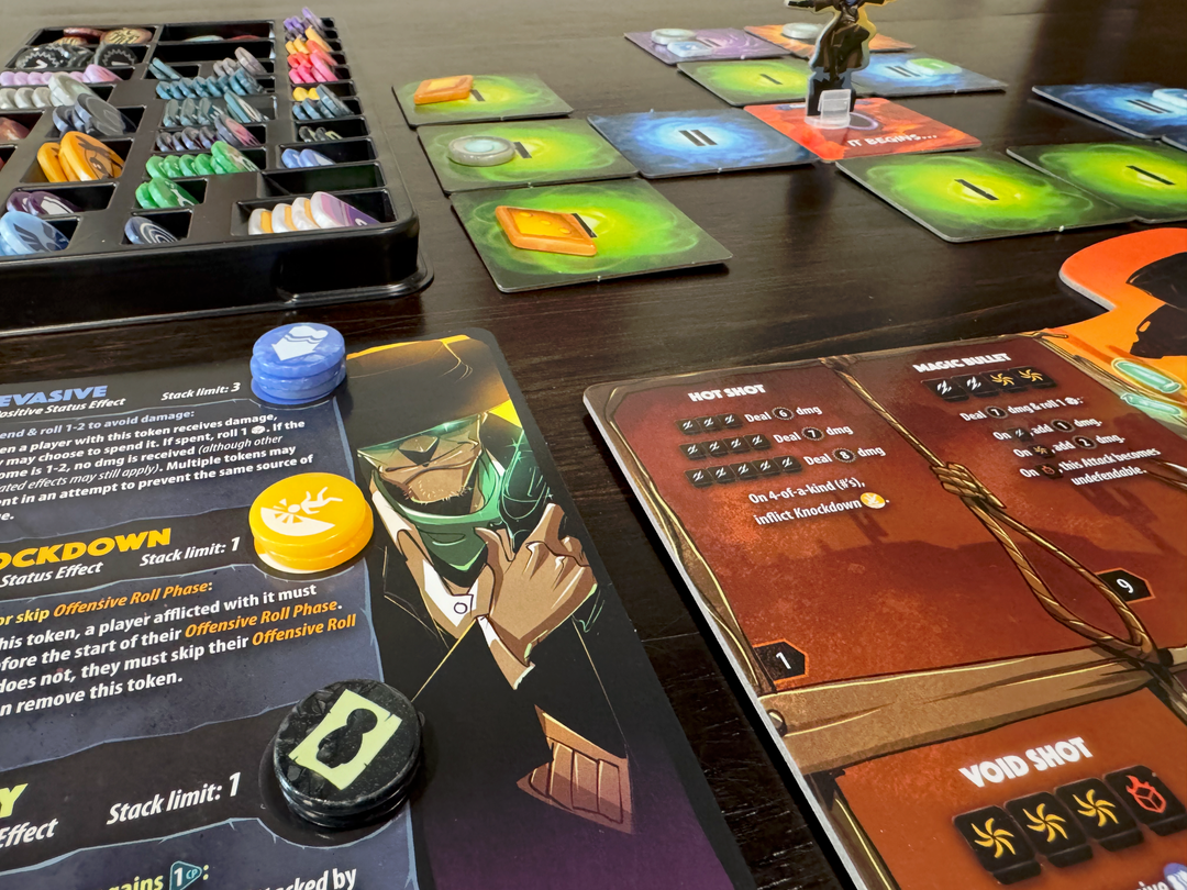 A photo of some of the original and upgraded pieces from the board game Dice Throne Adventures, pictured on a dark, wooden tabletop. A character set with text, an illustration of a man in a hat, and plastic GeekUp tokens is visible in the front. Tiles from the game are set up in the background with GeekUp bits on top of some of the tiles. In the background is also a compartmentalized tray with the rest of the GeekUp bits stored within,