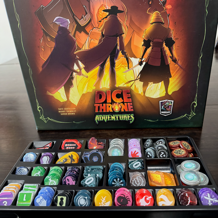 A picture of the GeekUp bits for use with the board game Dice Throne Adventures. All of the upgraded plastic bits are stored within a black, plastic tray, with individual compartments for each type of piece.  Behind the tray is the cover for the game, featuring the game's title and artwork depicting three characters standing in from of a giant, glowing, fourth figure.