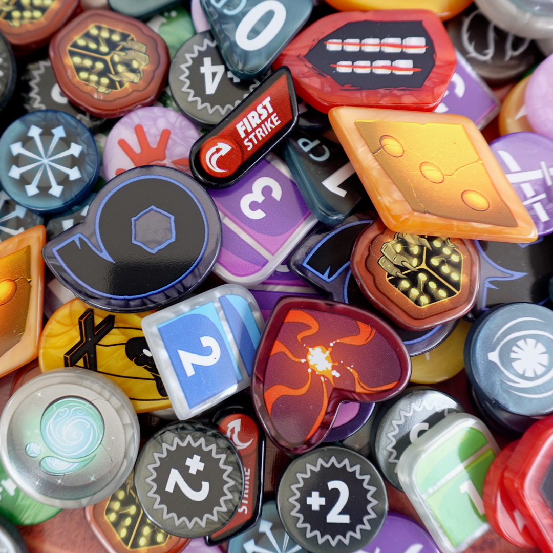 A photo of GeekUp bits for use with the board game Dice Throne Adventures. Each piece is made from pearlescence plastic and has an image from the game on both sides. The pieces are dumped in a pile on top of one of the player boards from the game.