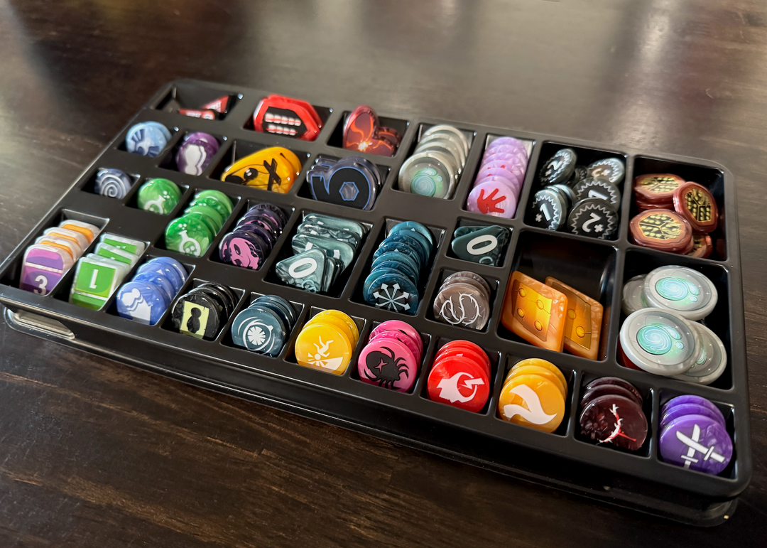 A photo of the GeekUp bits for use with the board game Dice Throne Adventures. All of the upgraded plastic bits are stored within a black, plastic tray, with individual compartments for each type of piece.   The tray is sitting on top of a glossy, dark wood tabletop.