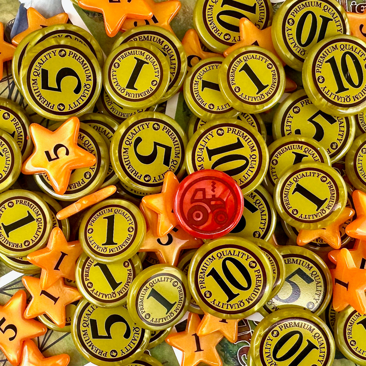 An overhead photo of a jumble of plastic GeekUp bits for use with the board game Fields of Green. Some bits are circular coins made from pearlescent gold plastic with the values of 1, 5 or 10. Other bits are orange five-pointed stars with the values 1 or 5. A single circular token is red with an outline of a tractor.