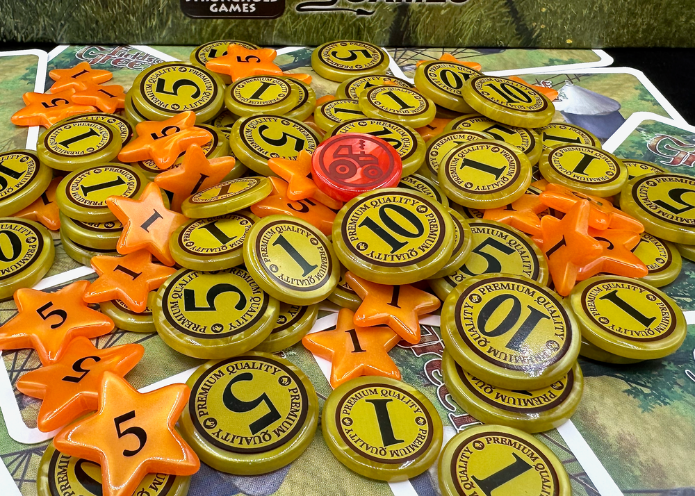 A side-view photo of a jumble of plastic GeekUp bits for use with the board game Fields of Green. Some bits are circular coins made from pearlescent gold plastic with the values of 1, 5 or 10. Other bits are orange five-pointed stars with the values 1 or 5. A single circular token is red with an outline of a tractor.