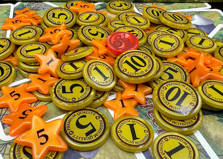 A side-view photo of a jumble of plastic GeekUp bits for use with the board game Fields of Green. Some bits are circular coins made from pearlescent gold plastic with the values of 1, 5 or 10. Other bits are orange five-pointed stars with the values 1 or 5. A single circular token is red with an outline of a tractor.