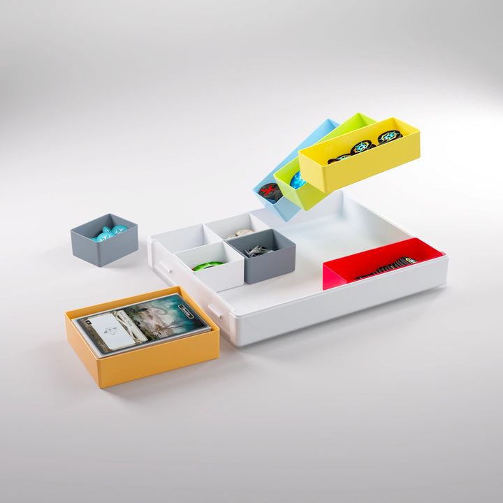 A photo of a white, plastic container on a white background. The plastic container has smaller, multicolored containers, some inside the larger frame, some sitting to the side, and some floating above. All of the smaller containers are filled with board game pieces and cards.