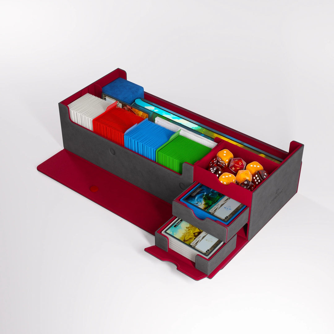 A photo of the gray/red version of the Cards' Lair Pro, made by Gamegenic, featuring a row of cards stacked upright, two drawer extended, a container with a variety of dice, and a long compartment containing a rolled mat.