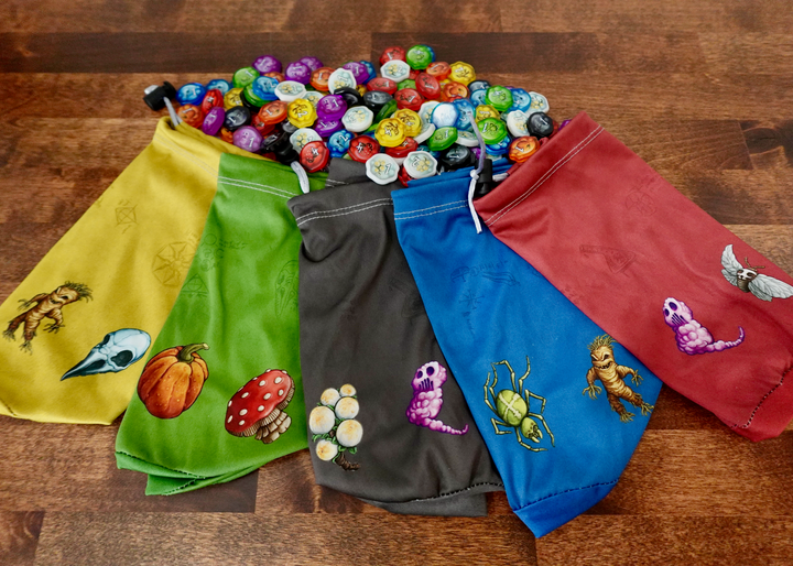 A set of microfiber drawstring bags for use with the board game Quacks of Quedlinburg, each with different colored background and potion ingredient symbols along the bottom, like a pumpkin, mushroom, spider, mandrake, and moth.  