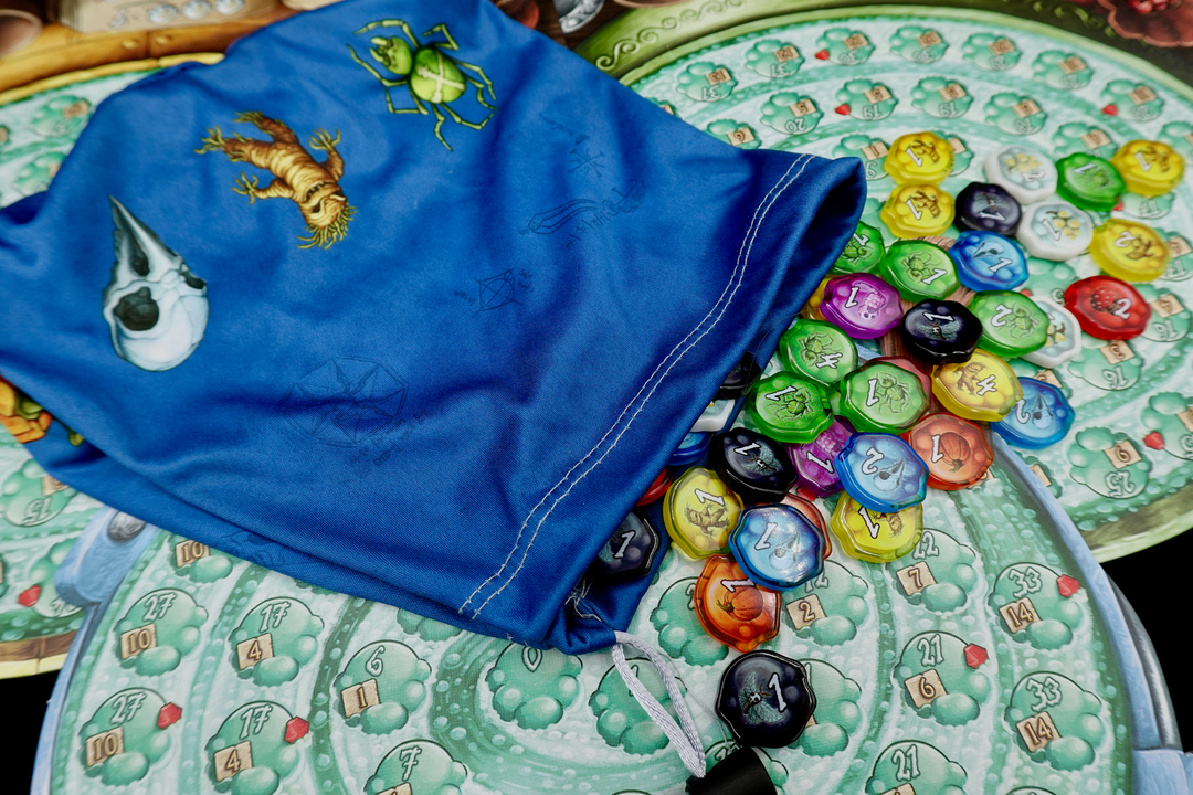 A single, microfiber drawstring bag for use with the board game Quacks of Quedlinburg, with a blue background a colorful symbols of skull, mandrake, and spider along the bottom. The bag is resting on player boards and a pile of plastic upgraded token for the same game. 