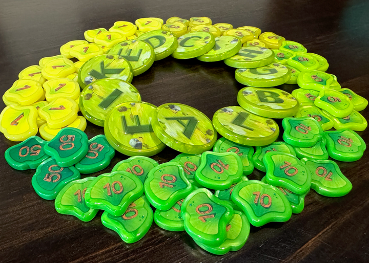 A photo of GeekUp tokens for use with the board game Ginkgopolis. A ring of circular tokens is in the center of the photo, with a single letter printed on each token. Surrounding those lettered tokens are are variety of smaller pieces, each in the shape of a Ginko leaf and each printed with a number in the center. All tokens are sitting on a glossy, dark wood tabletop.